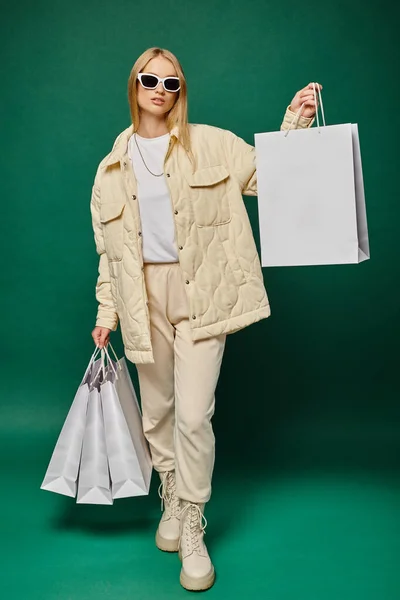 Donna bionda alla moda in abiti invernali e occhiali da sole in posa con borse della spesa su sfondo verde — Foto stock