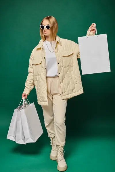 Giovane donna bionda in abbigliamento invernale e occhiali da sole in piedi con borse della spesa su sfondo verde — Foto stock