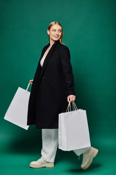 Pleine longueur de femme blonde joyeuse en manteau chaud noir marchant avec des sacs à provisions blancs sur vert — Photo de stock