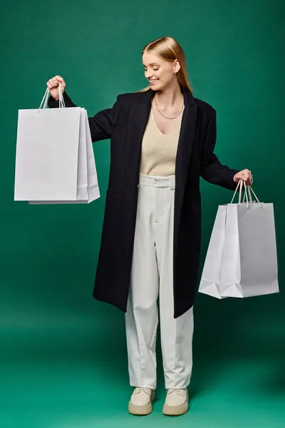 Full length of pleased blonde woman in black warm coat standing with white shopping  bags on green — Stock Photo