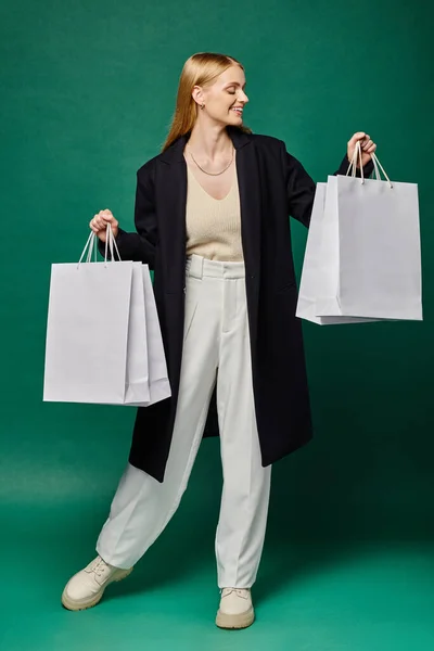 Delighted blonde woman in black stylish coat with white shopping  bags on green, winter sales — Stock Photo
