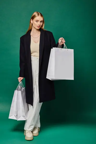 Longitud completa de la mujer rubia con estilo en el abrigo negro posando con bolsas de compras blancas en verde - foto de stock
