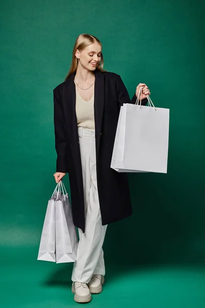 Piena lunghezza di donna bionda sorridente in caldo cappotto nero guardando borse della spesa bianche sul verde — Stock Photo