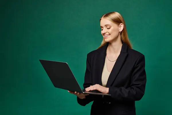 Glückliche blonde Frau im schwarzen Wintermantel mit Laptop auf grünem Hintergrund, freiberuflich und modisch — Stockfoto