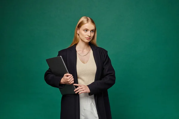 Femme blonde à la mode en manteau d'hiver noir tenant ordinateur portable et regardant loin sur fond vert — Photo de stock