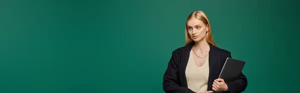 Fashionable blonde woman in black winter coat standing with laptop on green backdrop, banner — Stock Photo