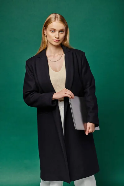 Young blonde woman in black winter coat holding laptop and looking at camera on green backdrop — Stock Photo