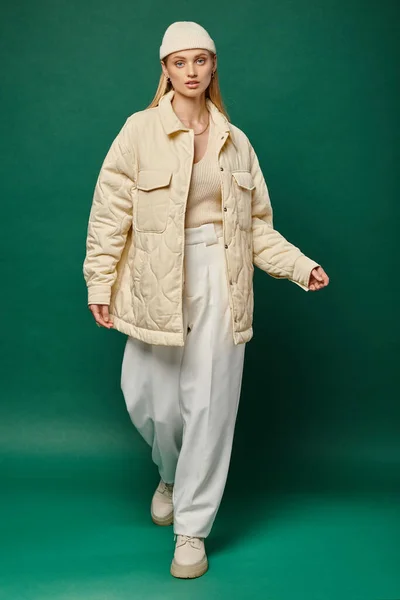 Modello femminile affascinante e alla moda in giacca avorio e cappello beanie che cammina sul verde, moda invernale — Foto stock