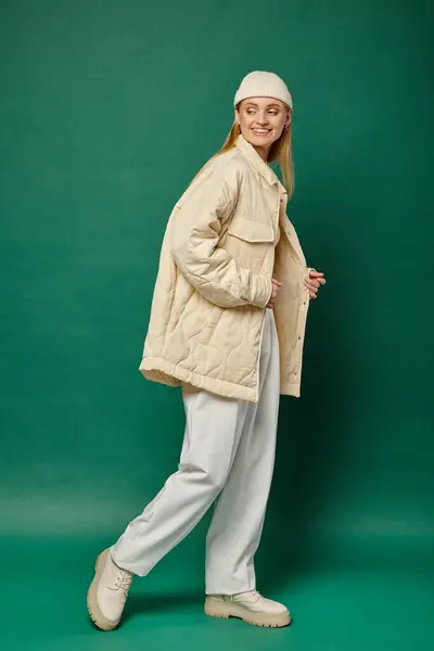 Счастливая и модная модель в пиджаке из слоновой кости и шапочке шапочка, идущая по зеленой, зимней моде — стоковое фото