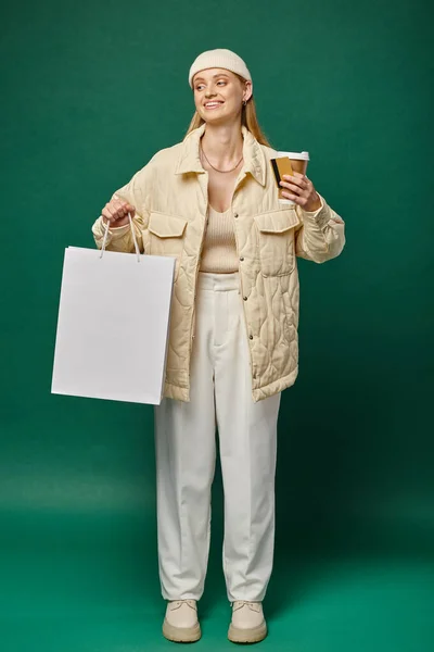 Mujer feliz en ropa de invierno con estilo con bolsa de compras, bebida para llevar y tarjeta de crédito en verde - foto de stock