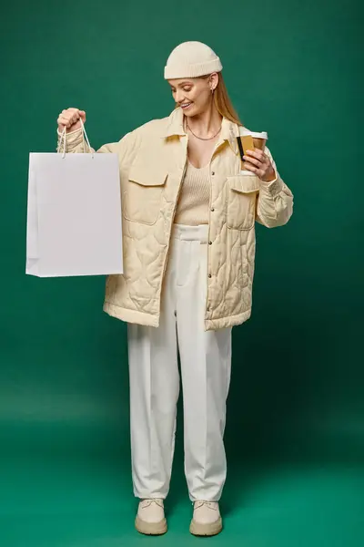 Веселая женщина в модном зимнем наряде с сумкой для покупок, горячим напитком и кредиткой на грине — стоковое фото