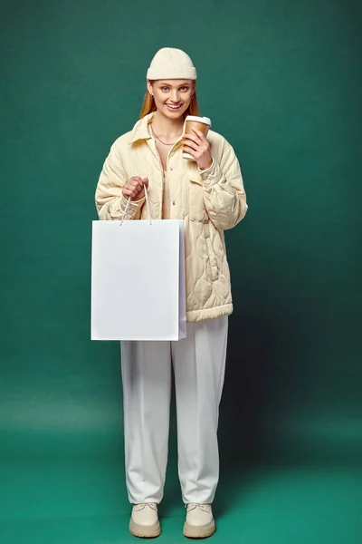Довольная молодая женщина в стильном зимнем наряде с сумкой для покупок и горячим напитком в бумажной чашке на зеленом — стоковое фото
