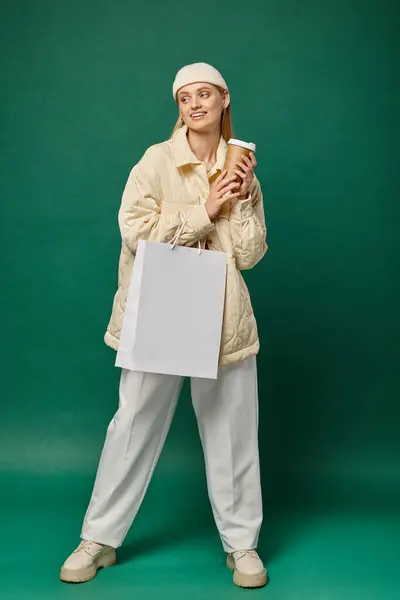 Young smiling woman in trendy winter clothing with shopping bag and coffee to go on green backdrop — Stock Photo