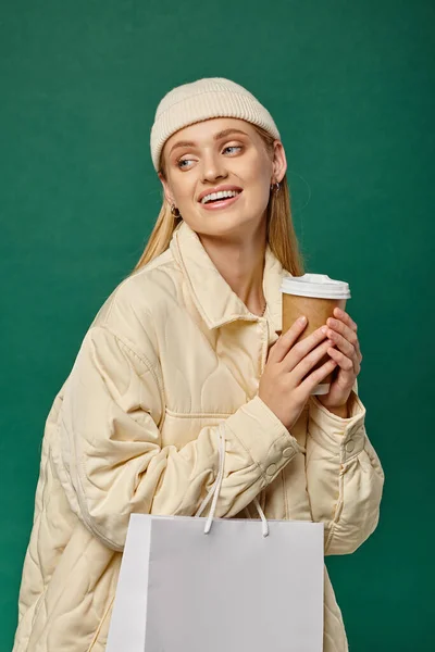 Беззаботная женщина в модной зимней куртке и шапочке с сумкой для покупок и горячим напитком на зеленом — стоковое фото