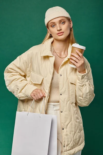 Trendy woman in warm winter clothes with shopping bag and coffee to go looking at on green — Stock Photo