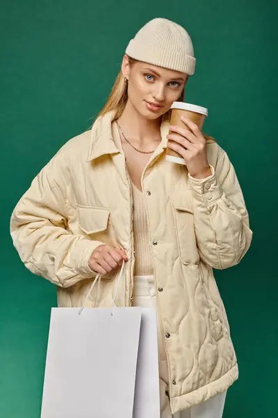 Stylish woman in cozy winter clothes with shopping bag and takeaway drink looking at camera on green — Stock Photo