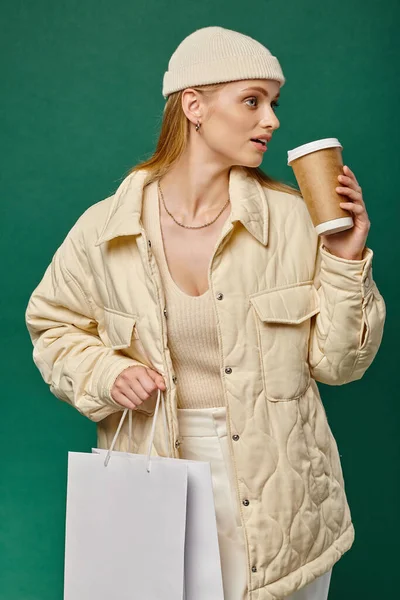 Young woman in stylish winter clothes with shopping bag and takeaway drink looking away on green — Stock Photo