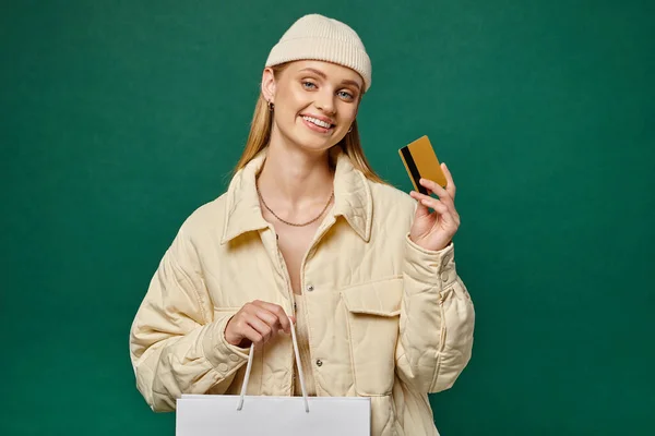 Щаслива жінка в стильному зимовому одязі з сумкою і кредитною карткою на зелених, зимових розпродажах — стокове фото