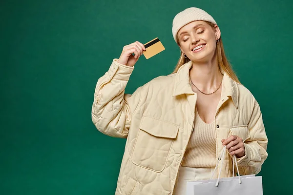 Mujer alegre con los ojos cerrados en traje de invierno de moda con bolsa de compras y tarjeta de crédito en verde - foto de stock