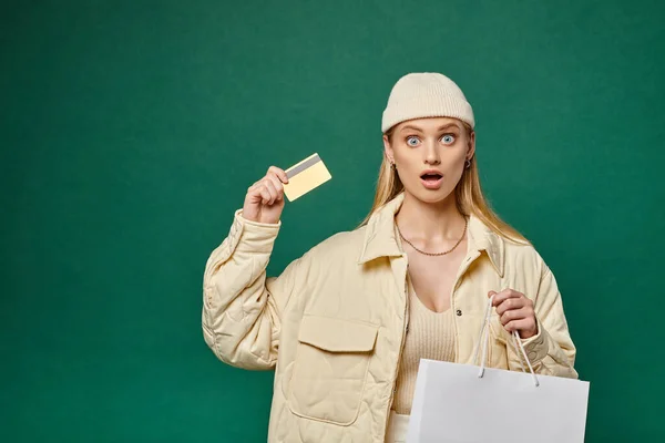 Amazed woman in stylish winter attire with shopping bag and credit card looking at camera on green — Stock Photo