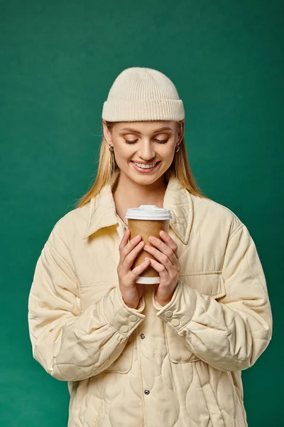 Pleased woman in beanie hat and warm trendy jacket holding hot takeaway drink on green, winter joy — Stock Photo