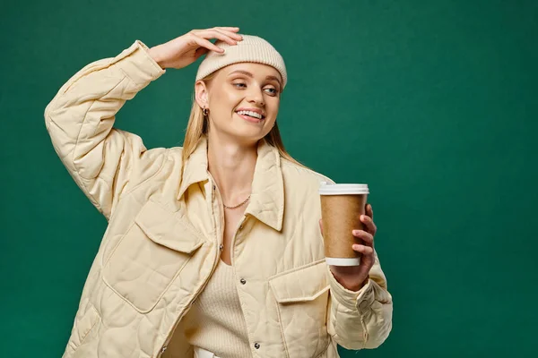 Cheerful woman in beanie hat and warm jacket holding hot takeaway drink and looking away on green — Stock Photo