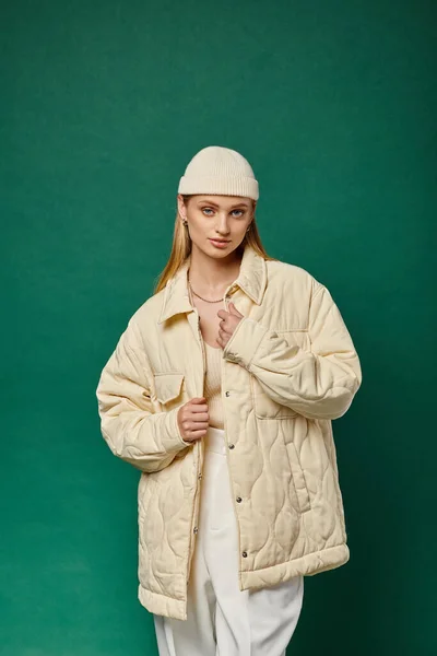 Young woman in warm ivory jacket and stylish beanie hat on green backdrop, winter fashion — Stock Photo