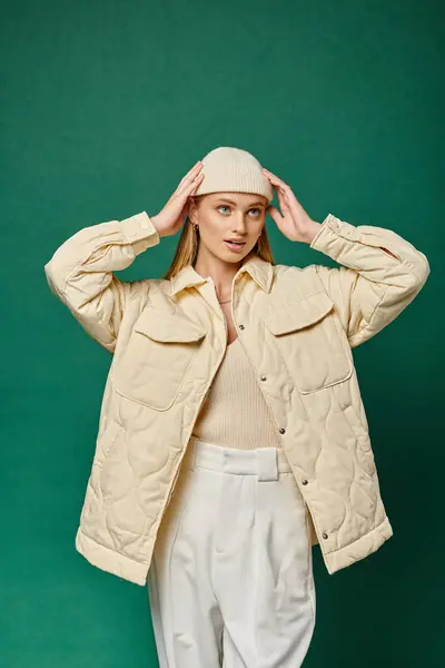 Mujer con estilo en chaqueta de marfil caliente ajustando gorro sombrero sobre fondo verde, moda casual de invierno - foto de stock