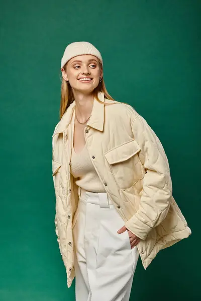 Mujer alegre en chaqueta elegante y sombrero de gorro mirando hacia otro lado en verde, emociones positivas de invierno - foto de stock