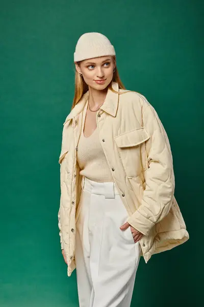Trendy woman in winter jacket and beanie hat posing with hand in pocket of white pants on green — Stock Photo