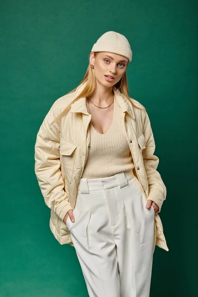Stylish woman in winter jacket and beanie hat posing with hands in pockets of white pants on green — Stock Photo