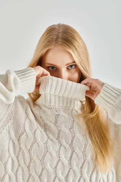 Stylische blonde Frau verschleiert Gesicht mit hohem Kragen aus weißem Strickpullover auf grauem, winterlichen Trend — Stockfoto