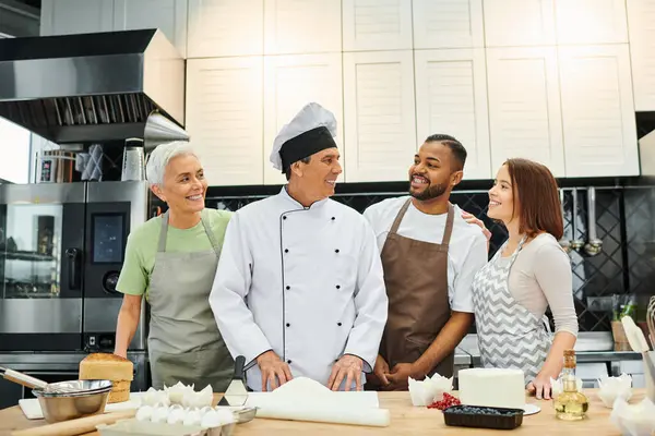 Красивый радостный шеф-повар смотрит на своих разнообразных учеников и счастливо улыбается во время урока кулинарии — стоковое фото