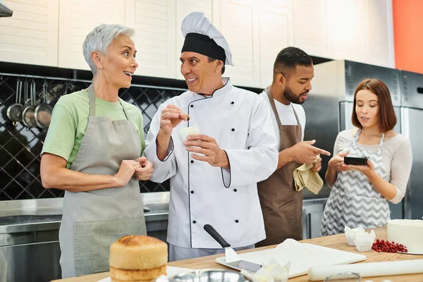 Allegro cuoco maturo in cappello bianco parlando con diversi studenti durante la lezione di cucina, pasticceria — Foto stock