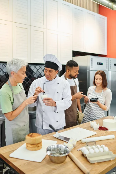 Persone multiculturali in grembiuli ascoltando maturo bel cuoco in cappello bianco, corsi di cucina — Foto stock