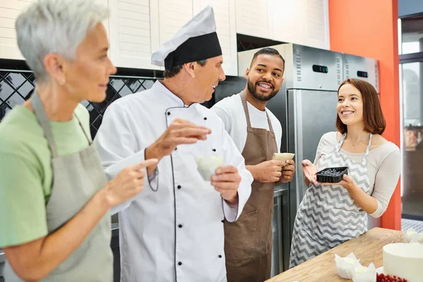 Gente multicultural alegre en delantales hablando con cocinero maduro en sombrero blanco, cursos de cocina - foto de stock