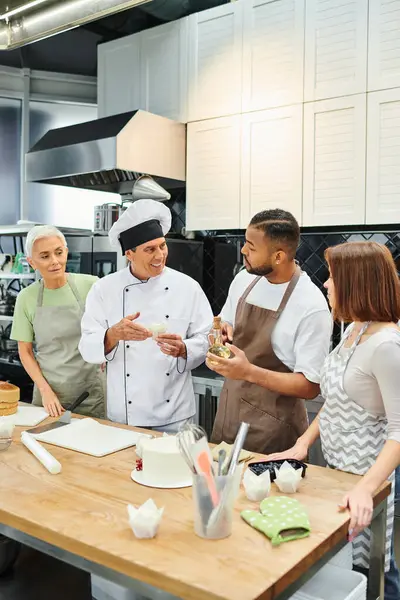 Alegre maduro chef en blanco sombrero hablando con su interracial estudiantes durante la cocina lección - foto de stock
