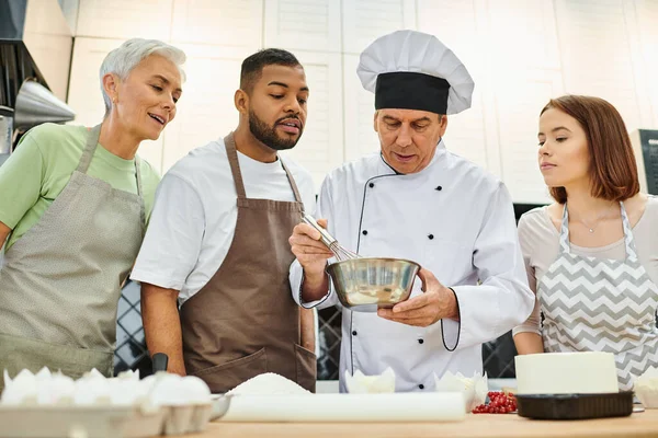 Handsome mature chef in white hat showing how to whisk dough to diverse students, cooking courses — Stock Photo