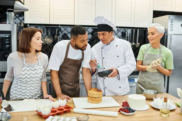 Estudiantes multirraciales guapos en delantales escuchando al chef en sombrero blanco durante la lección de cocina - foto de stock