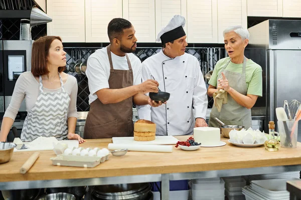 Estudiantes interraciales y chef en sombrero blanco mirando a la mujer madura haciendo preguntas, cursos de cocina - foto de stock
