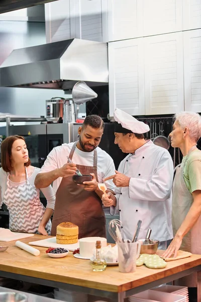 Alegre afroamericano hombre cepillado pastel con jarabe durante la lección junto a chef y sus amigos - foto de stock