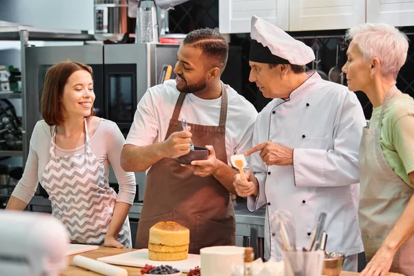 Jolly young woman looking at her multiracial friends and chef, student brushing cake with syrup — Stock Photo