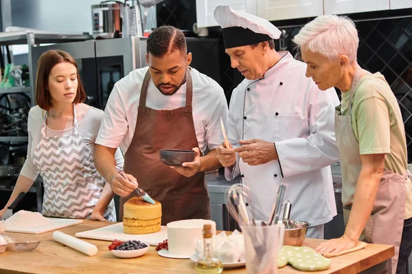 Estudiantes femeninas y chef maduro viendo hombre afroamericano cepillando pastel con jarabe en pastel - foto de stock