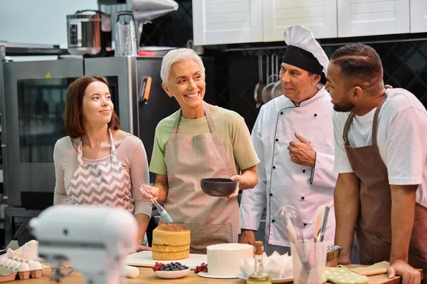 Cheerful mature woman with silicone brush smiling at her diverse friends and chef in white hat — Stock Photo