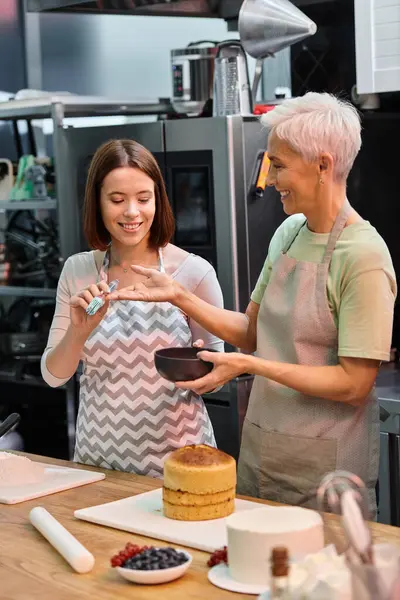 Mujer joven dando cepillo de silicona a su amigo alegre madura para usarlo en la torta, cursos de cocina - foto de stock