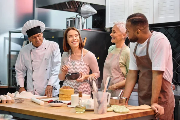 Joyeuse jeune femme brossant gâteau au sirop et souriant à ses amis divers à côté du chef mature — Photo de stock