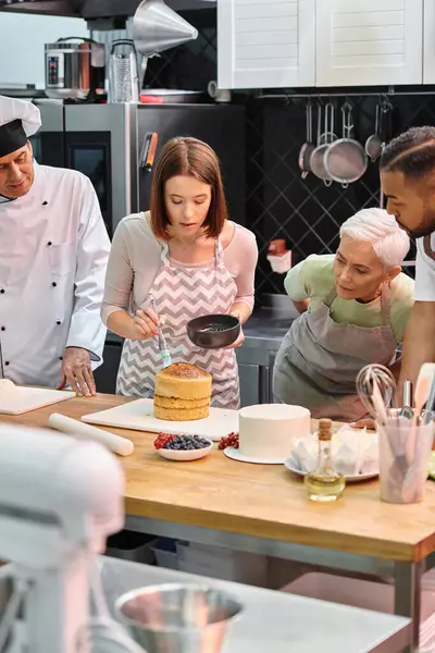Attrayant femme avec tablier brossage gâteau avec sirop sur gâteau à côté de ses divers amis et chef — Photo de stock
