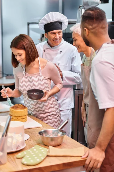 Giovane donna allegra spazzolatura torta con sciroppo mentre i suoi diversi amici e chef parlando attivamente — Foto stock