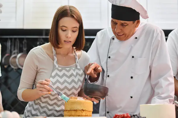 Веселий зрілий шеф-кухар в білому капелюсі навчає свою студентку, як правильно використовувати силіконову щітку — стокове фото