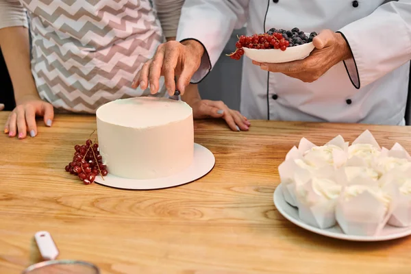 Chef mature montrant à son jeune étudiant comment décorer un gâteau savoureux avec des baies, cours de cuisine — Photo de stock